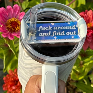 May include: A white and gray marble-patterned tumbler with a white handle. The lid has a clear straw and a blue rectangular accent piece with the text "fuck around and find out" in white script.
