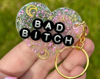 May include: A heart-shaped keychain with the words "BAD BITCH" in black and white letters. The keychain is clear with glitter and gold accents, including crescent moons and stars, and has a gold keyring.