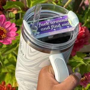 May include: A white and gray swirl-patterned tumbler with a clear lid and a clear straw. The lid has a purple and white accent piece with the text "fuck around and find out". The tumbler has a white handle.
