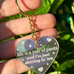 Peut inclure: Un porte-clés en forme de cœur iridescent avec une chaîne et un anneau dorés. Le porte-clés porte l'inscription "life is a pair of pants and I'm just shitting in 'em".