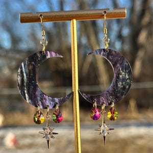 May include: A pair of celestial-themed earrings. Each earring features a crescent moon shape with a swirling purple and black pattern, adorned with dangling iridescent teardrop-shaped gems and a star pendant. The earrings are suspended from gold-colored hooks.