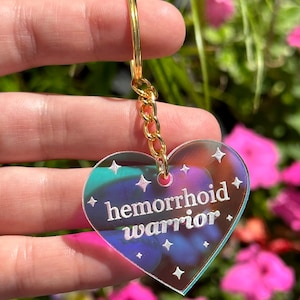May include: A heart-shaped iridescent keychain with a gold chain and the text "hemorrhoid warrior" in white.