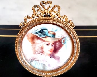 May include: A gold-toned metal frame with a decorative bow at the top holds a round porcelain portrait of a woman wearing a large hat with a blue ribbon. The portrait is painted in watercolor and has a delicate, vintage feel.