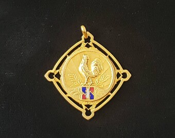 May include: A gold pendant featuring a detailed design of a rooster standing on a shield with the French flag colors. The pendant is surrounded by a decorative, openwork frame.