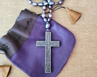 May include: A silver cross pendant with a pearl and iridescent stone inlay hangs from a long strand of dark purple pearls. The necklace is displayed on a purple velvet pouch.