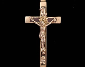 May include: A silver and gold crucifix pendant with a dark brown background. The crucifix features a figure of Jesus Christ on the cross with a skull and crossbones at the bottom.