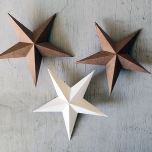 以下が含まれることがあります： 薄い木製の表面に、茶色の星が2つと白い星が1つ、合計3つの紙製の星が並んでいます。星はすべて5つの角を持ち、シンプルで折り畳まれたデザインです。
