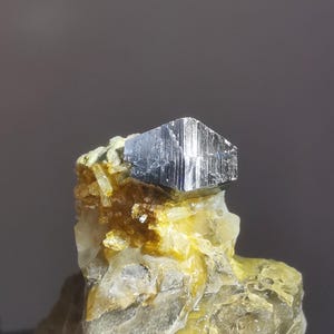 以下が含まれることがあります： 幾何学的な長方形の形をした、銀色の金属鉱物標本。鉱物は、黄色と白の結晶質の岩の母岩にセットされています。背景は無地のニュートラルグレーです。