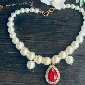K&ouml;nnte beinhalten: Eine wei&szlig;e Perlenkette mit einer goldenen Kette und einem roten Edelstein-Anh&auml;nger. Der Anh&auml;nger hat die Form einer Tr&auml;ne und ist von kleineren, klaren Edelsteinen umgeben.