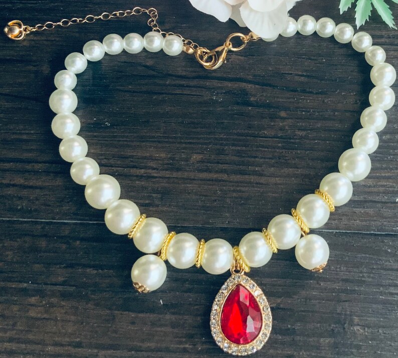 K&ouml;nnte beinhalten: Eine wei&szlig;e Perlenkette mit einer goldenen Kette und einem roten Edelstein-Anh&auml;nger. Der Anh&auml;nger hat die Form einer Tr&auml;ne und ist von kleineren, klaren Edelsteinen umgeben.