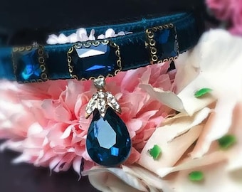 Puede incluir: Un collar de terciopelo azul oscuro para gato con un gran colgante de cristal azul en forma de lágrima. El collar está decorado con cristales azules más pequeños de forma cuadrada.
