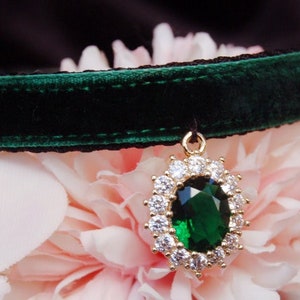 May include: A dark green velvet choker with a gold pendant featuring a large emerald green gemstone surrounded by smaller clear gemstones.