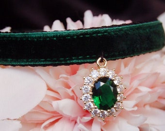 May include: A dark green velvet choker with a gold pendant featuring a large emerald green gemstone surrounded by smaller clear gemstones.