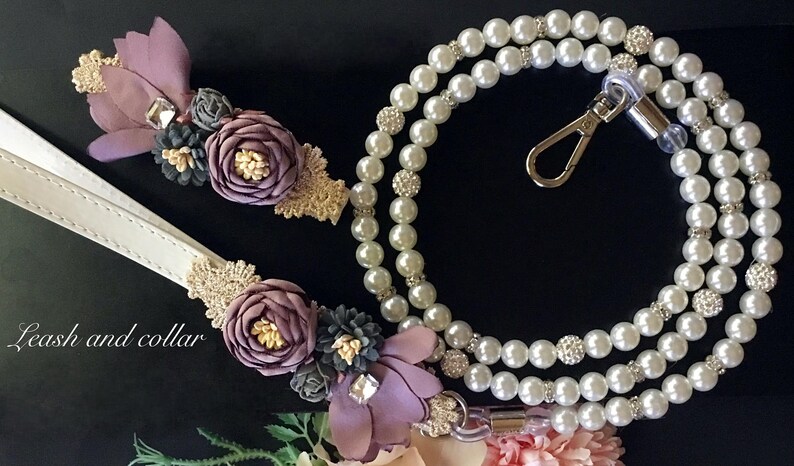 May include: A white leather leash and a pearl necklace with a silver clasp. Both are adorned with purple, gold, and gray fabric flowers.