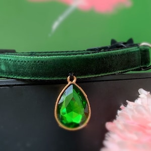May include: A dark green velvet cat collar with a gold-tone pendant featuring a large, faceted, emerald green gemstone.