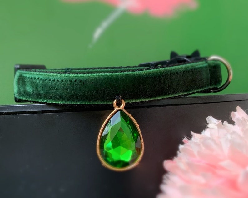 May include: A dark green velvet cat collar with a gold-tone pendant featuring a large, faceted, emerald green gemstone.