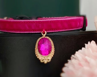 May include: A pink velvet cat collar with a gold-tone pendant featuring a large, pink gem.