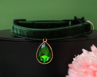 May include: A dark green velvet cat collar with a gold-tone pendant featuring a large, emerald green gemstone.
