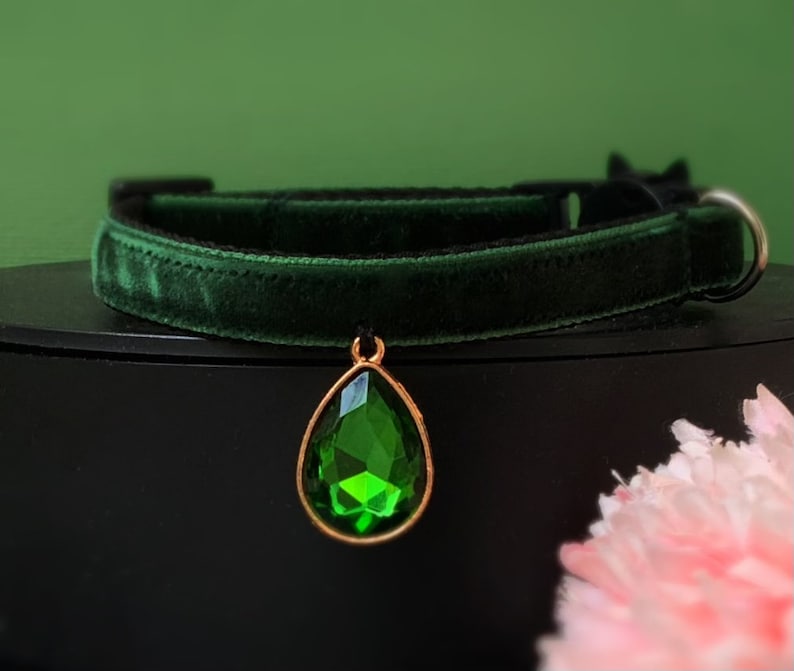 May include: A dark green velvet cat collar with a gold-tone pendant featuring a large, emerald green gemstone.