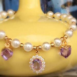 May include: A delicate bracelet made of white pearls with three dangling charms. Each charm features a purple gemstone surrounded by gold accents.