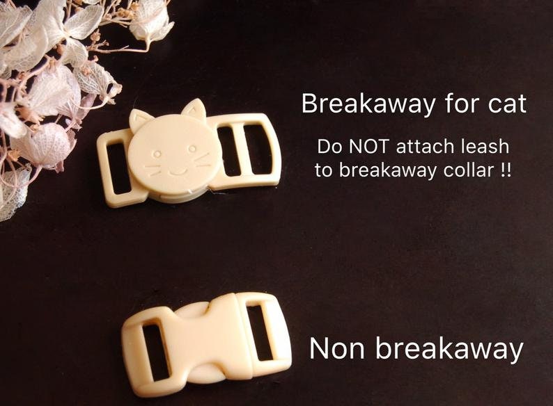 May include: Two beige plastic cat collar buckles. The top buckle has a cat face design and the text "Breakaway for cat. Do NOT attach leash to breakaway collar!" The bottom buckle has the text "Non breakaway".