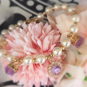 May include: A delicate pearl bracelet with a gold chain and two purple gemstone charms. The bracelet is displayed on a pink flower.