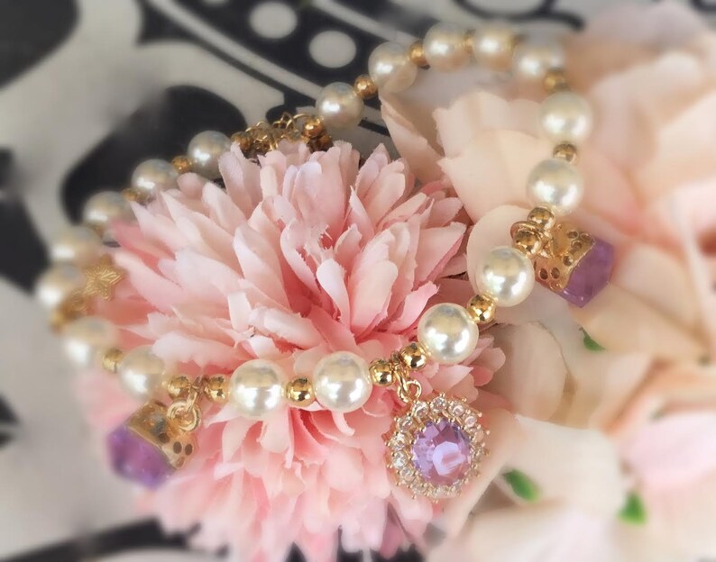 May include: A delicate pearl bracelet with a gold chain and two purple gemstone charms. The bracelet is displayed on a pink flower.