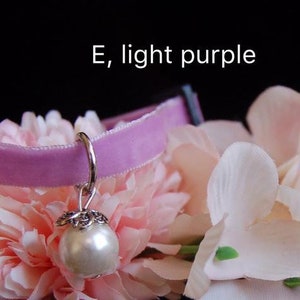 May include: A light purple velvet choker with a silver charm featuring a pearl. The choker is displayed on a bed of pink flowers.
