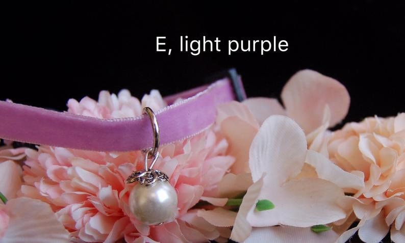 May include: A light purple velvet choker with a silver charm featuring a pearl. The choker is displayed on a bed of pink flowers.