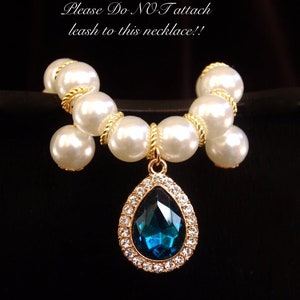 K&ouml;nnte beinhalten: Eine wei&szlig;e Perlenkette mit einer goldenen Kette und einem tropfenf&ouml;rmigen Anh&auml;nger mit einem blauen Edelstein, der von klaren Strasssteinen umgeben ist. Die Kette tr&auml;gt ein Schild mit der Aufschrift "Bitte KEINE Leine an diese Halskette befestigen!"