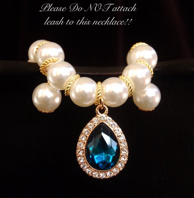 K&ouml;nnte beinhalten: Eine wei&szlig;e Perlenkette mit einer goldenen Kette und einem tropfenf&ouml;rmigen Anh&auml;nger mit einem blauen Edelstein, der von klaren Strasssteinen umgeben ist. Die Kette tr&auml;gt ein Schild mit der Aufschrift "Bitte KEINE Leine an diese Halskette befestigen!"