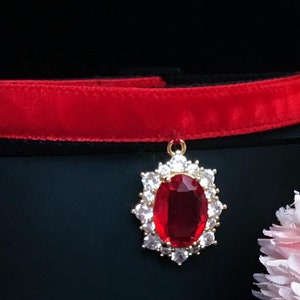 May include: A red velvet choker with a gold pendant featuring a large red gemstone surrounded by smaller clear gemstones.