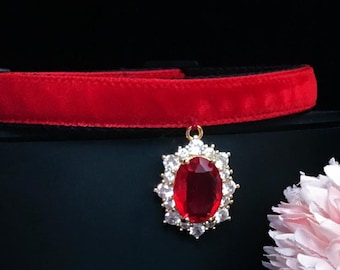 Puede incluir: Un collar gargantilla de terciopelo rojo con un colgante dorado que presenta una gran piedra roja rodeada de piedras transparentes más pequeñas.