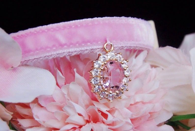 May include: A pink velvet cat collar with a gold pendant featuring a large oval-shaped pink gemstone surrounded by clear rhinestones.