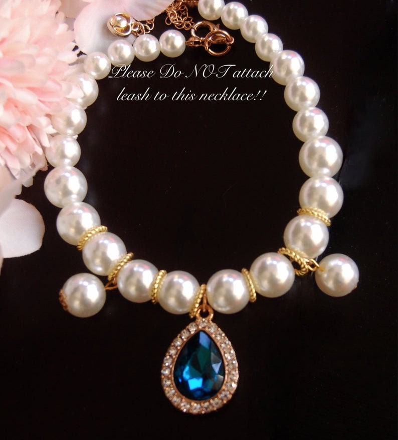 K&ouml;nnte beinhalten: Eine wei&szlig;e Perlenkette mit einer goldenen Kette und einem tropfenf&ouml;rmigen Anh&auml;nger mit einem blauen Edelstein. Die Kette tr&auml;gt ein Schild mit der Aufschrift "Bitte KEINE Leine an diese Kette befestigen!"