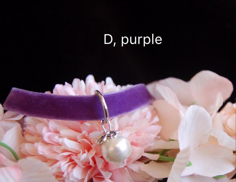 May include: Purple velvet choker with a silver charm and a white pearl pendant. The choker is displayed on a bed of pink flowers.  D, purple.