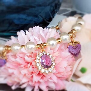 May include: A delicate pearl bracelet with two purple heart-shaped charms. The bracelet is adorned with small gold beads and is displayed on a pink flower.