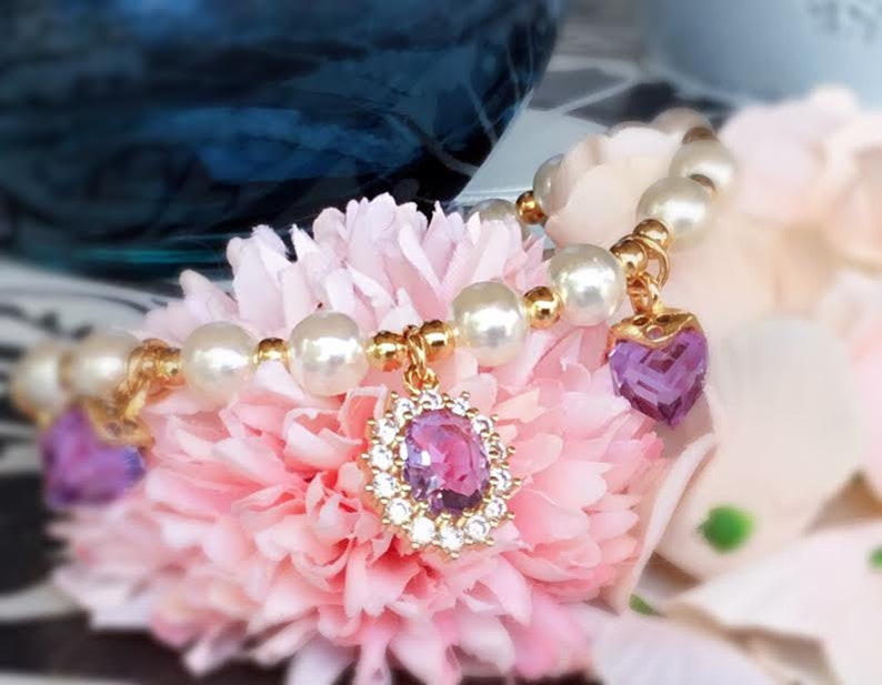 May include: A delicate pearl bracelet with two purple heart-shaped charms. The bracelet is adorned with small gold beads and is displayed on a pink flower.