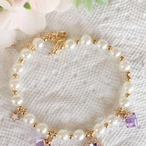 May include: A delicate pearl bracelet with gold accents and purple gemstone charms. The bracelet features a star charm, a heart charm, and a round charm with a floral design.