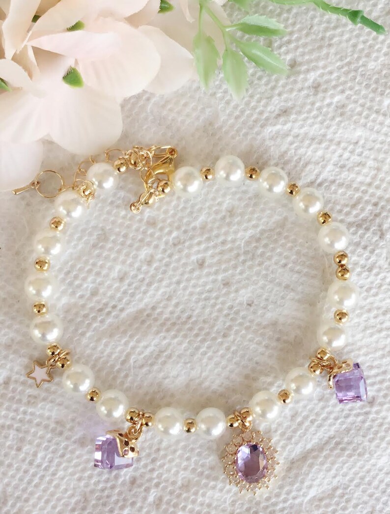 May include: A delicate pearl bracelet with gold accents and purple gemstone charms. The bracelet features a star charm, a heart charm, and a round charm with a floral design.