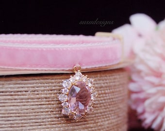 May include: A pink velvet cat collar with a gold charm featuring a large, clear pink gemstone surrounded by smaller clear gemstones.