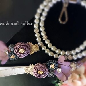 May include: A white leather dog collar and leash with a pearl and rhinestone chain. The collar and leash are decorated with purple and gold fabric flowers.