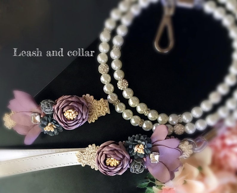 May include: A white leather dog collar and leash with a pearl and rhinestone chain. The collar and leash are decorated with purple and gold fabric flowers.