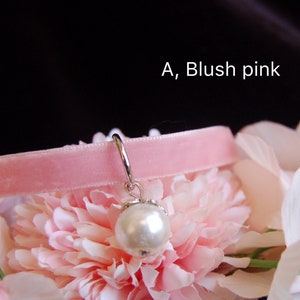May include: A blush pink velvet choker with a silver charm featuring a white pearl. The choker is displayed on a bed of pink and white flowers.