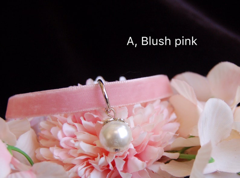 May include: A blush pink velvet choker with a silver charm featuring a white pearl. The choker is displayed on a bed of pink and white flowers.