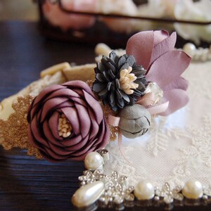 May include: A delicate floral hair accessory with a purple rose, gray flower, and a purple fabric flower. The accessory is adorned with a crystal and pearl accents.