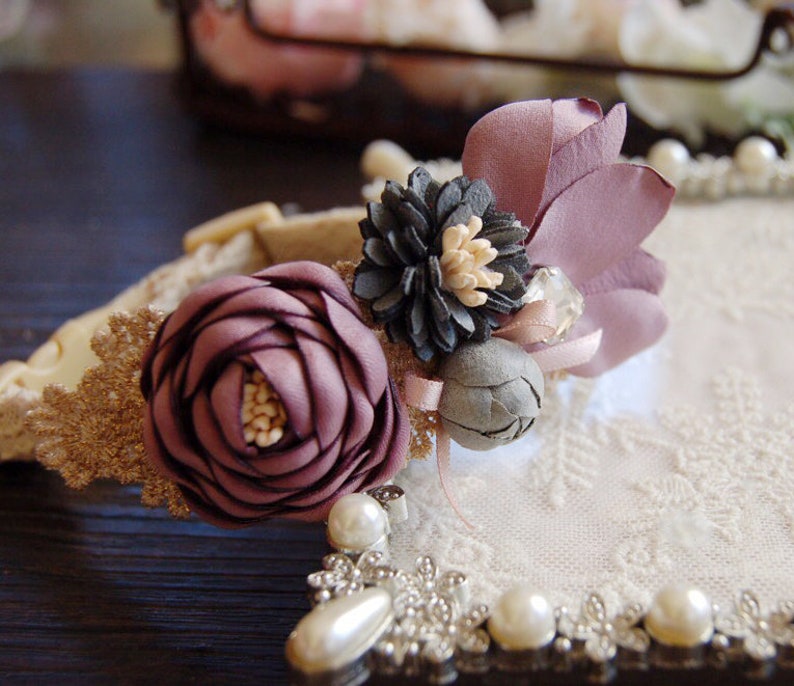 May include: A delicate floral hair accessory with a purple rose, gray flower, and a purple fabric flower. The accessory is adorned with a crystal and pearl accents.