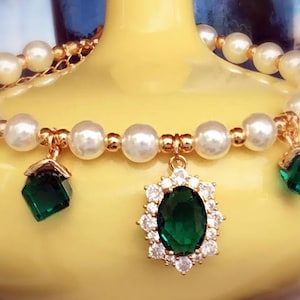 May include: A gold bracelet with white pearl beads and three green gemstone charms. The center charm is a large oval-shaped gemstone surrounded by smaller white gemstones. The other two charms are square-shaped.