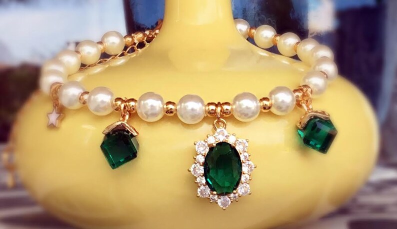 May include: A gold bracelet with white pearl beads and three green gemstone charms. The center charm is a large oval-shaped gemstone surrounded by smaller white gemstones. The other two charms are square-shaped.