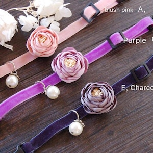 Può includere: Tre collari in velluto per animali domestici in rosa cipria, viola e grigio antracite. Ogni collare presenta un fiore in tessuto e un ciondolo a perla. I collari hanno fibbie in plastica nera e sono esposti su una superficie di legno.